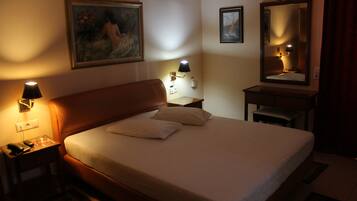 Una habitación de hotel con cama, mesitas de noche, lámpara, cuadro, espejo y escritorio.
