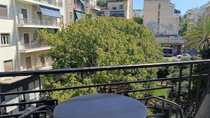 A balcony with a round table and chairs, overlooking a cityscape with multi-story buildings and a large tree. - Exis Boutique Hotel (Athens)