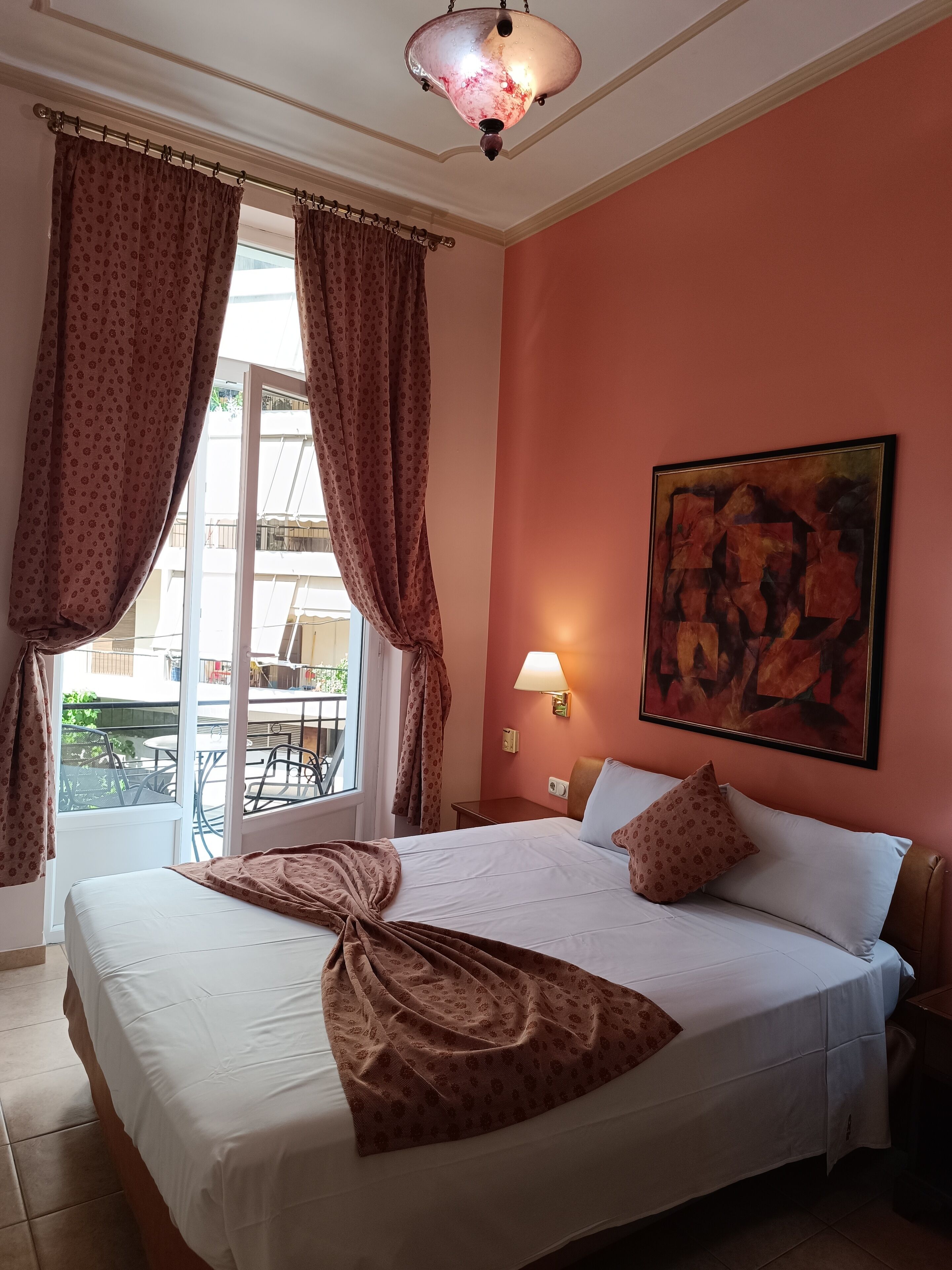 A hotel room with a bed, a painting on the wall, a window with curtains, and a view of the outside.