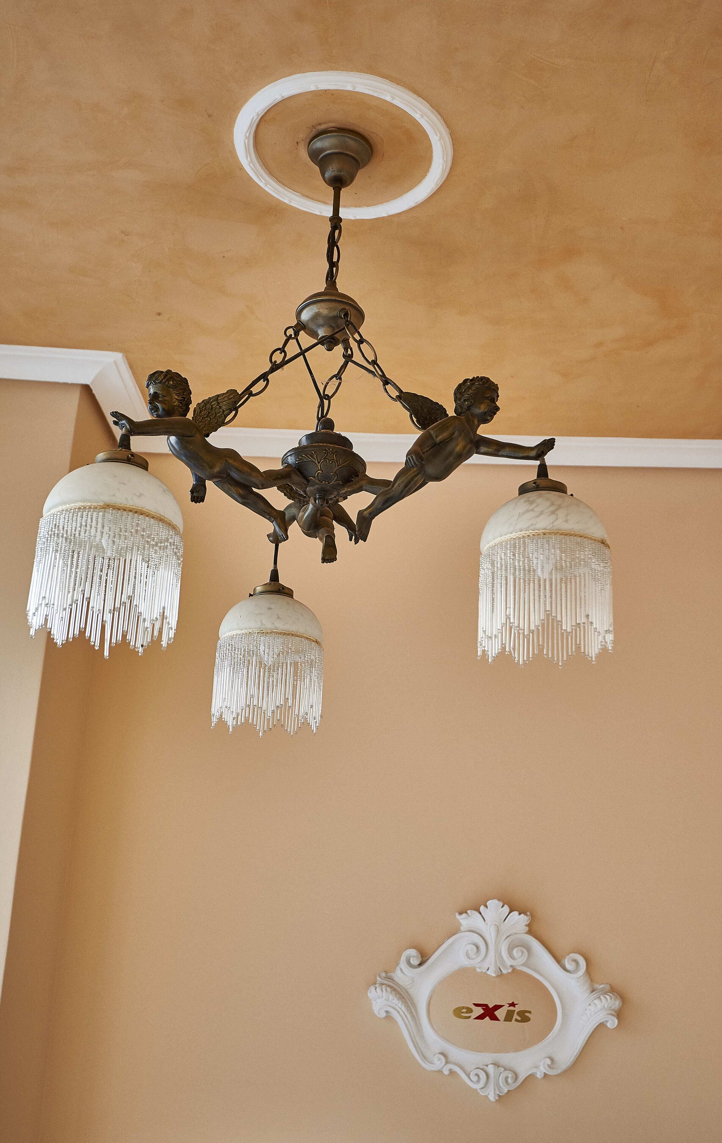 A chandelier with multiple arms and hanging fringes, a decorative ceiling medallion, and a framed picture with the text "eXis" on a beige wall.