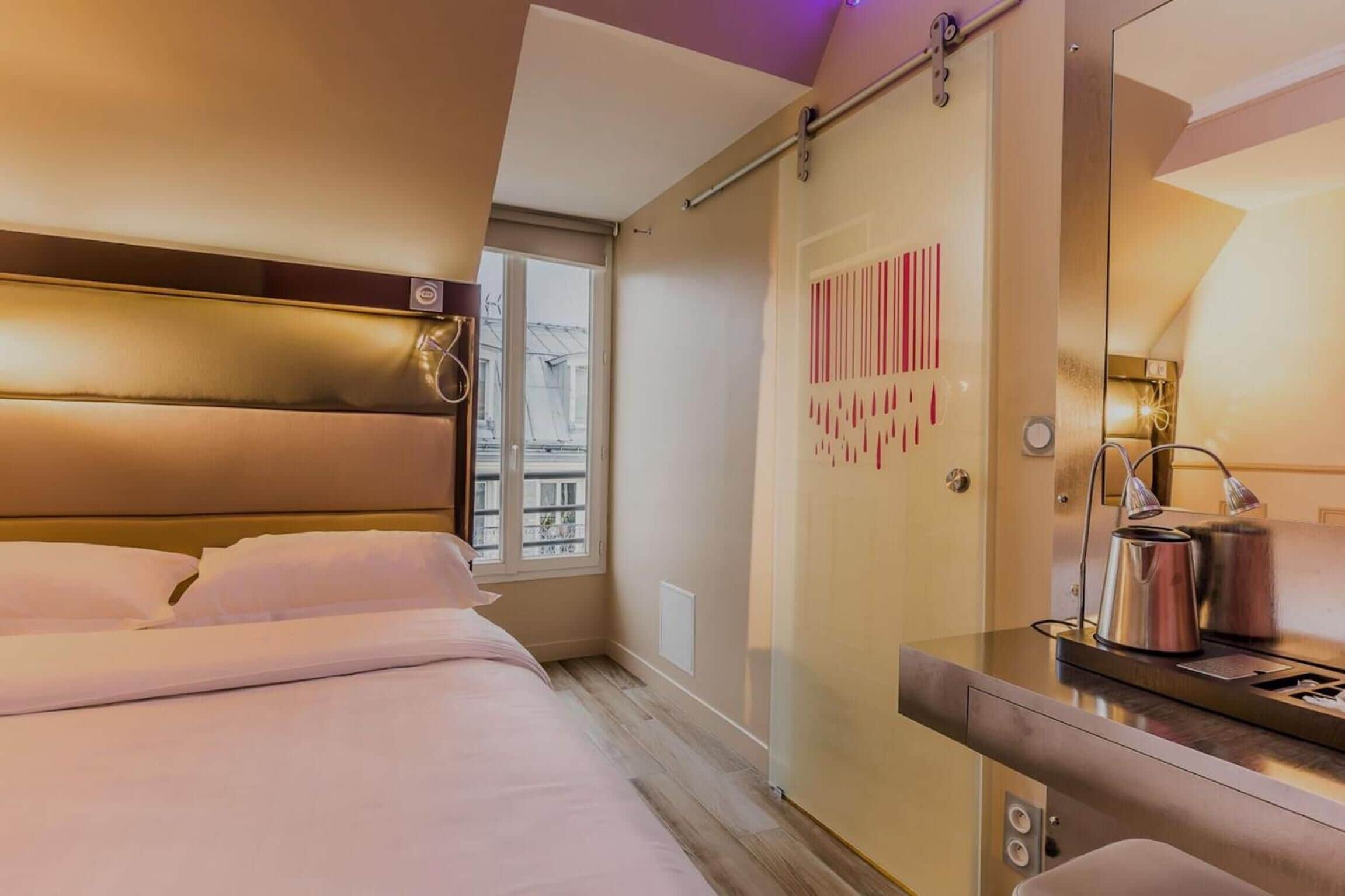 A hotel room with a bed, a bedside table with a kettle, a mirror, and a sliding door with a striped pattern.