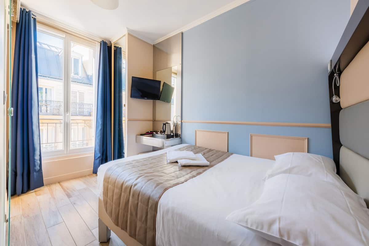 A hotel room with a bed, a TV, a bedside table, and a window with curtains.
