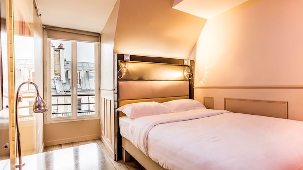 Quarto de hotel com cama, janela com vista para prédios e porta com porta-toalhas.