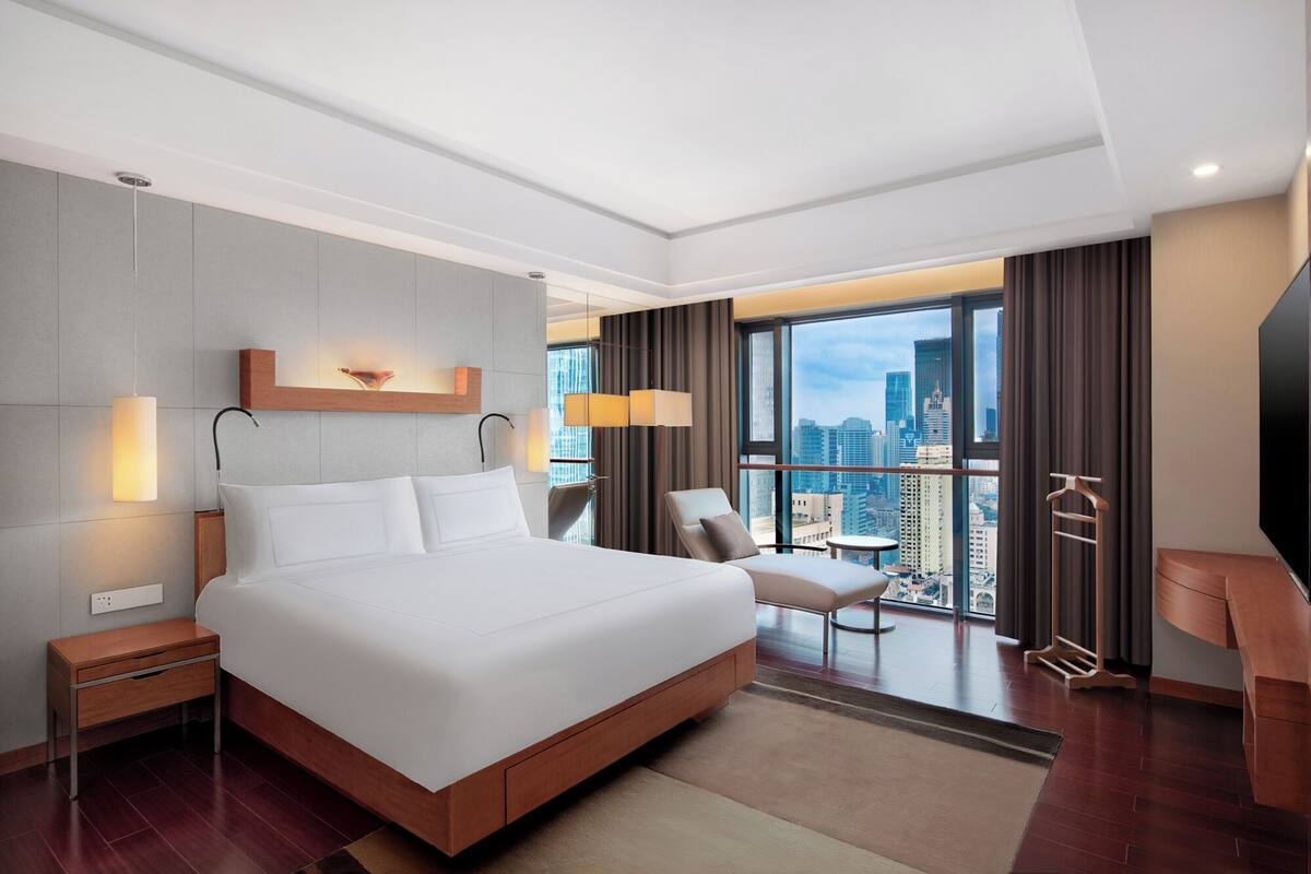 Quarto de hotel moderno com uma cama grande, vista para a cidade, escrivaninha e uma cadeira.