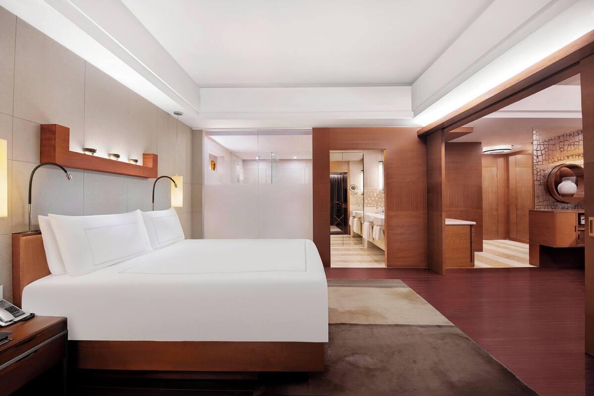 Quarto de hotel moderno com uma cama grande, uma mesa com um laptop e um banheiro visível por uma porta aberta.