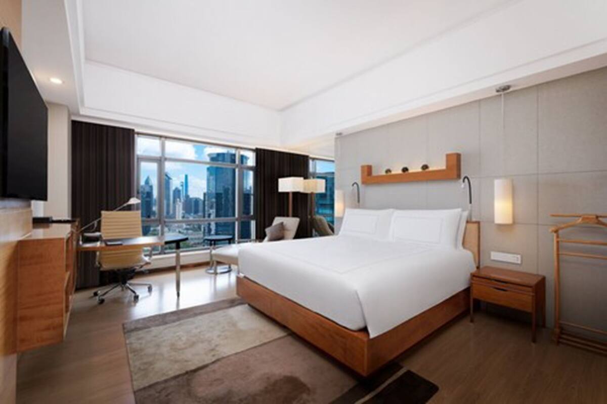 Quarto de hotel moderno com uma cama grande, uma escrivaninha, uma cadeira, um abajur e vista para a cidade.
