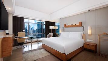 A modern hotel room with a large bed, a desk, a chair, a lamp, and a view of the city.