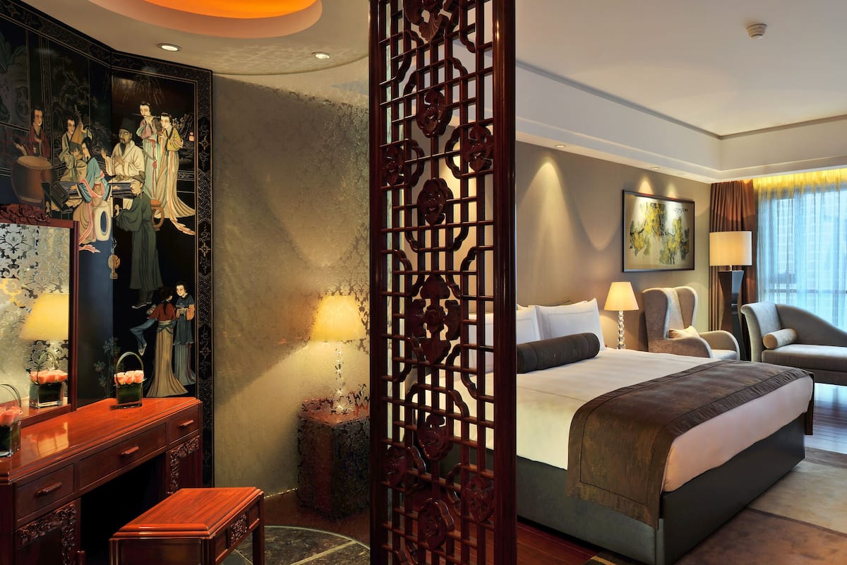 Quarto de hotel com uma cama grande, uma escrivaninha com candeeiro, uma cadeira e um ecrã decorativo.