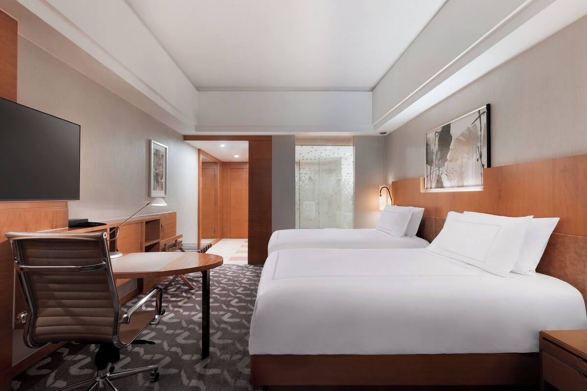 Quarto de hotel com cama, escrivaninha, cadeira, televisão e janela.