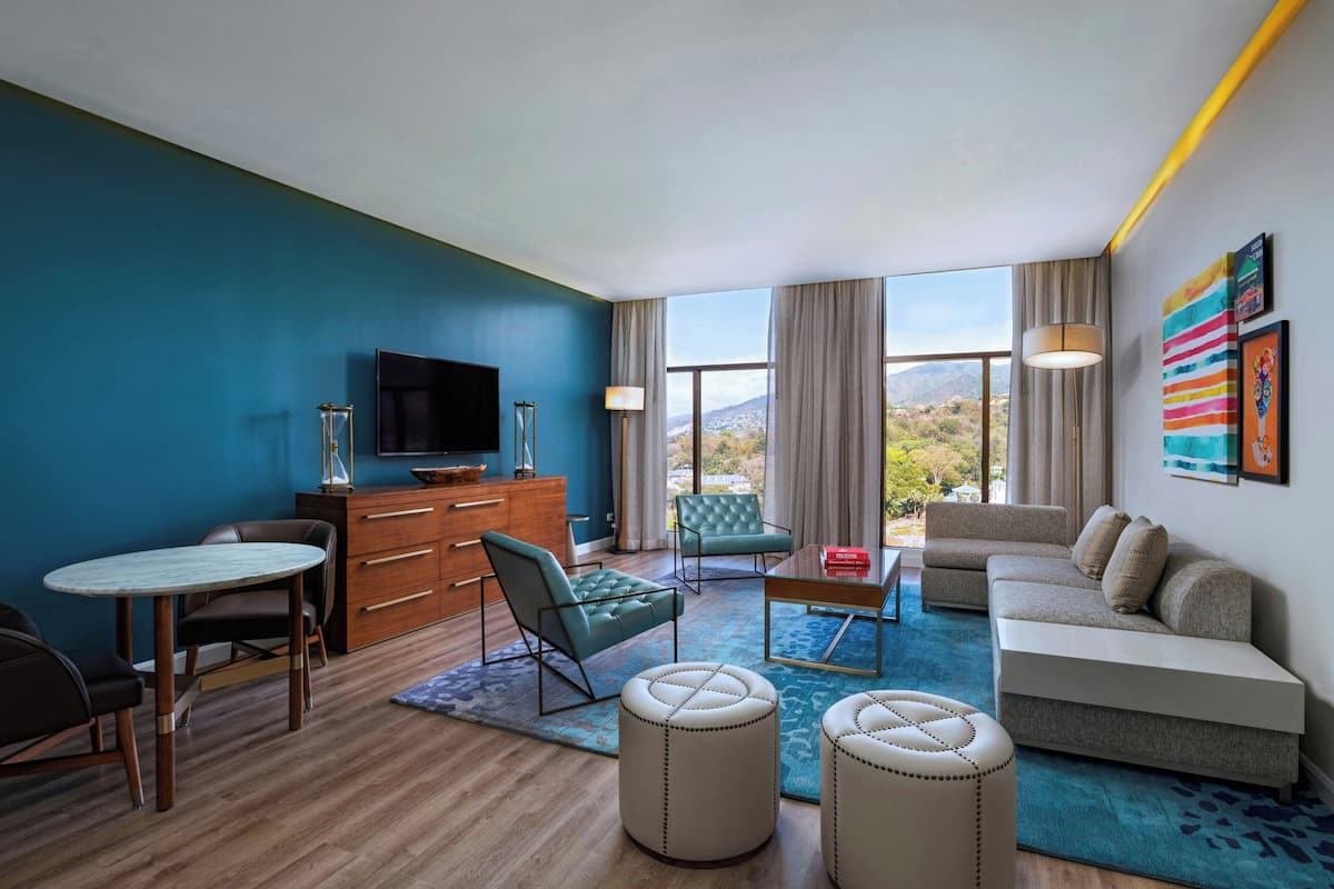 A modern hotel room with a blue accent wall, a wooden floor, a sofa, a TV, a dining table, and a view of the mountains.