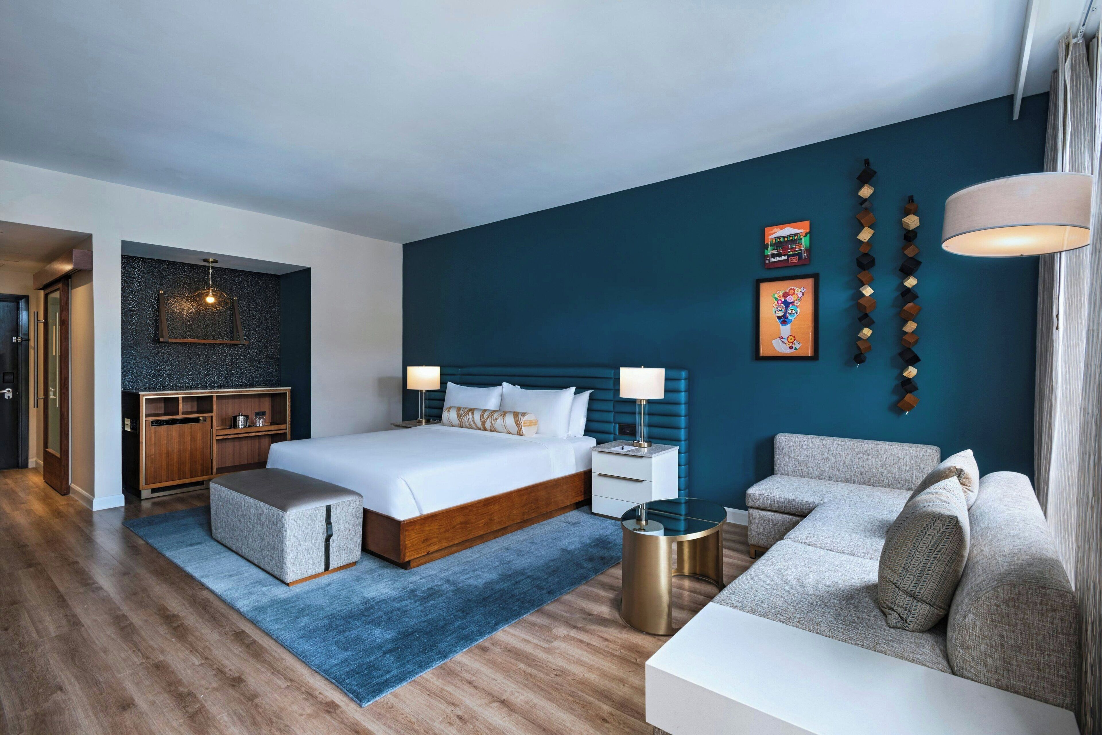 Una habitación de hotel moderna con una cama grande, un sofá, una mesita y un escritorio. Se observa un armario de madera y una lámpara colgante.