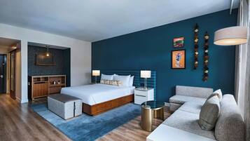 Una habitación de hotel moderna con una cama grande, un sofá, una mesita y un escritorio. Se observa un armario de madera y una lámpara colgante.