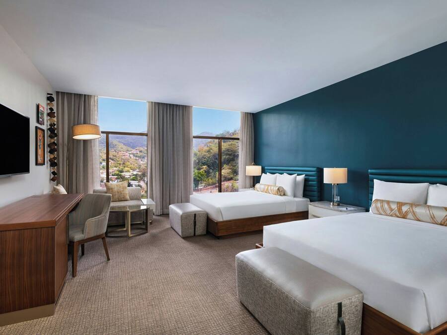 A hotel room with a large bed, a smaller bed, a desk, a chair, a TV, and a view of the outdoors.