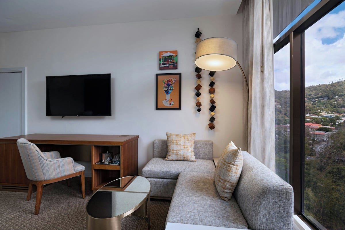 A modern living room with a sofa, armchair, and a glass coffee table. A flat-screen TV is mounted on the wall, and there are two framed artworks above the sofa.