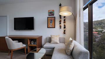 A modern living room with a sofa, armchair, and a glass coffee table. A flat-screen TV is mounted on the wall, and there are two framed artworks above the sofa.