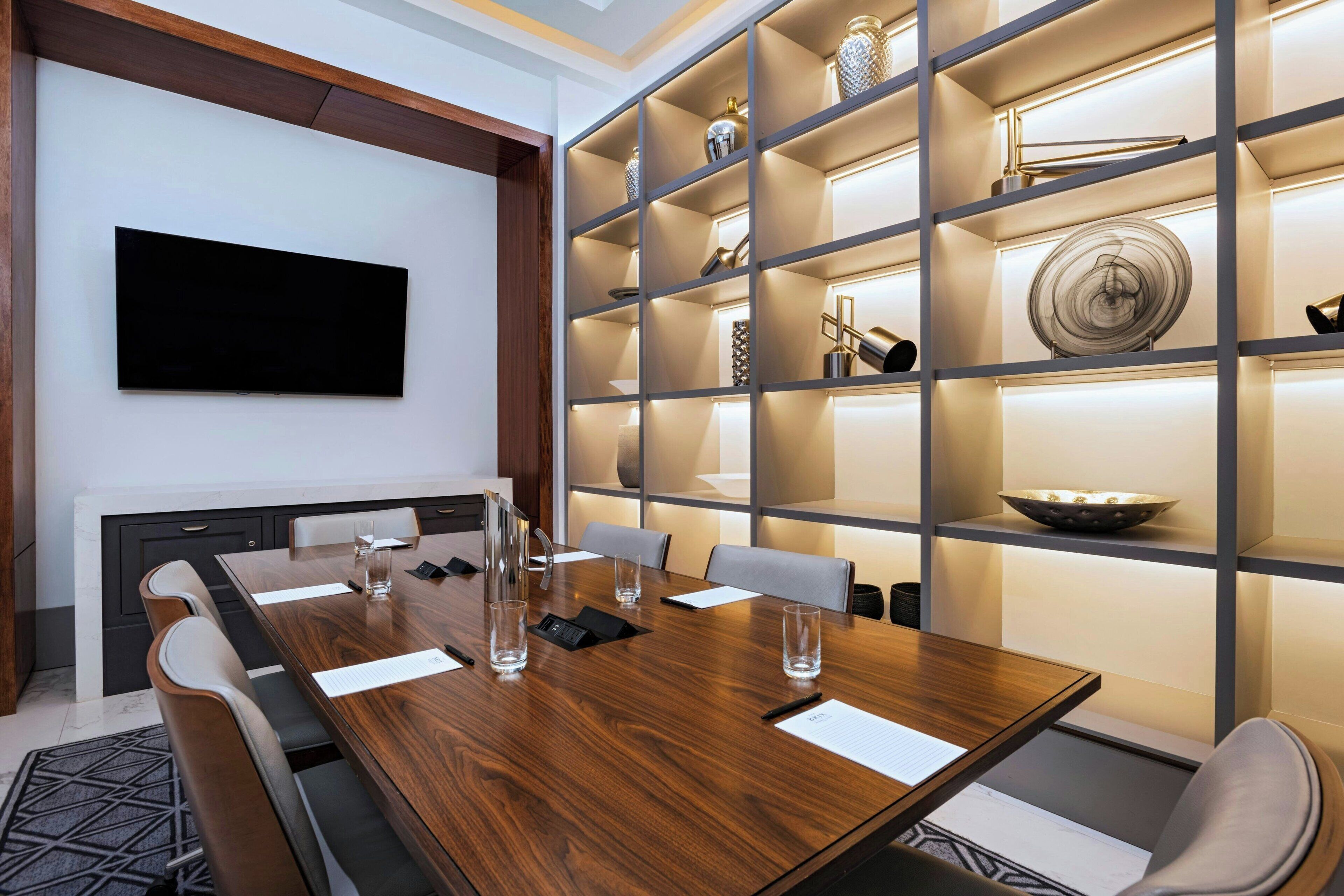 A conference room with a long wooden table, chairs, a flat-screen TV, and a shelving unit with decorative items.