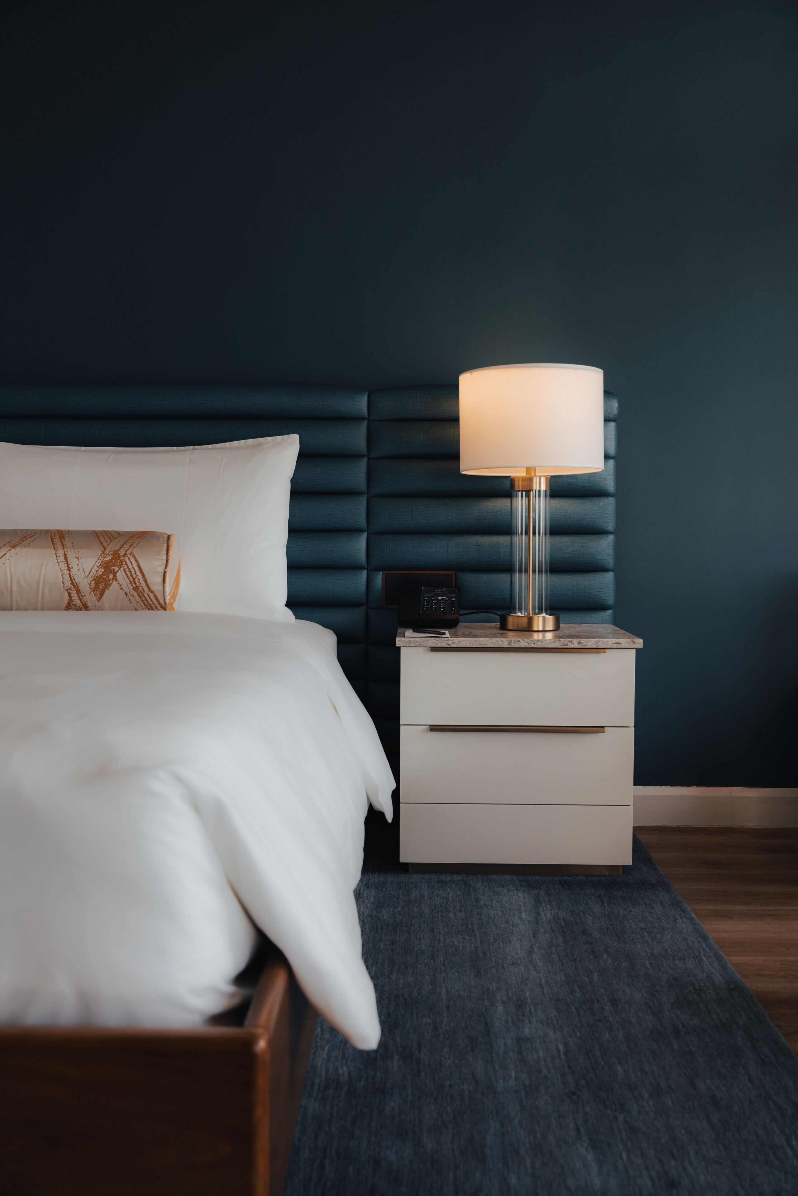 A neatly made bed with white linens, a bedside table with a lamp, and a dark blue upholstered headboard.