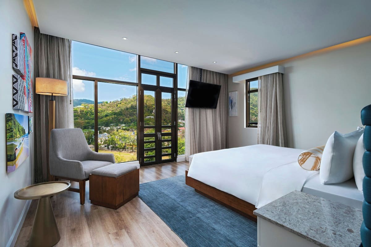 A modern hotel room with a large bed, a seating area, a flat-screen TV, and a view of a green landscape through large windows.