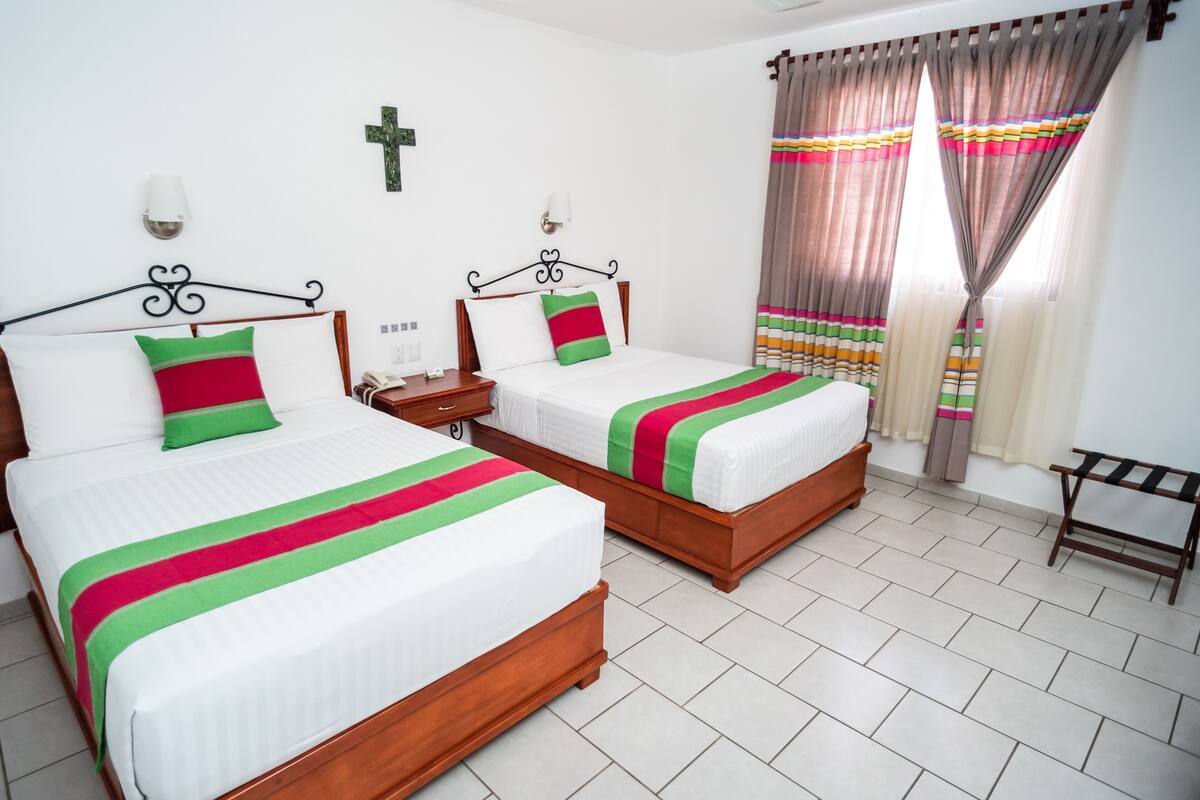 A hotel room with two beds, a nightstand, a cross on the wall, and curtains.