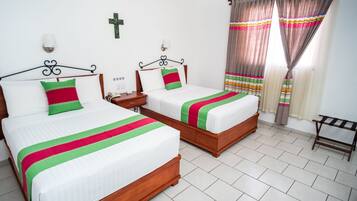 Habitación de hotel con dos camas, una mesita de noche, una cruz en la pared y cortinas.