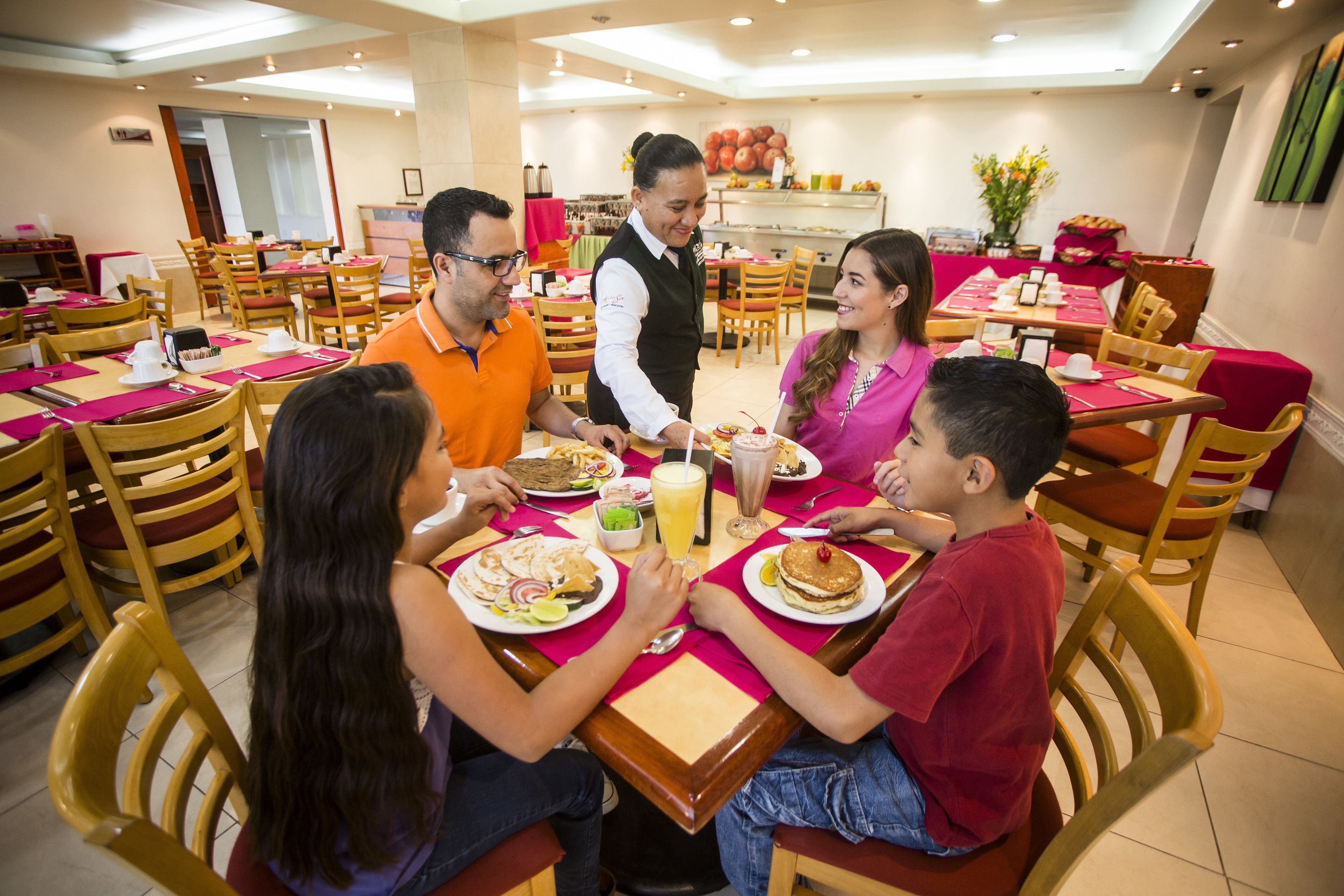 A dining area with wooden chairs and tables, a waiter serving food, and a buffet in the background.