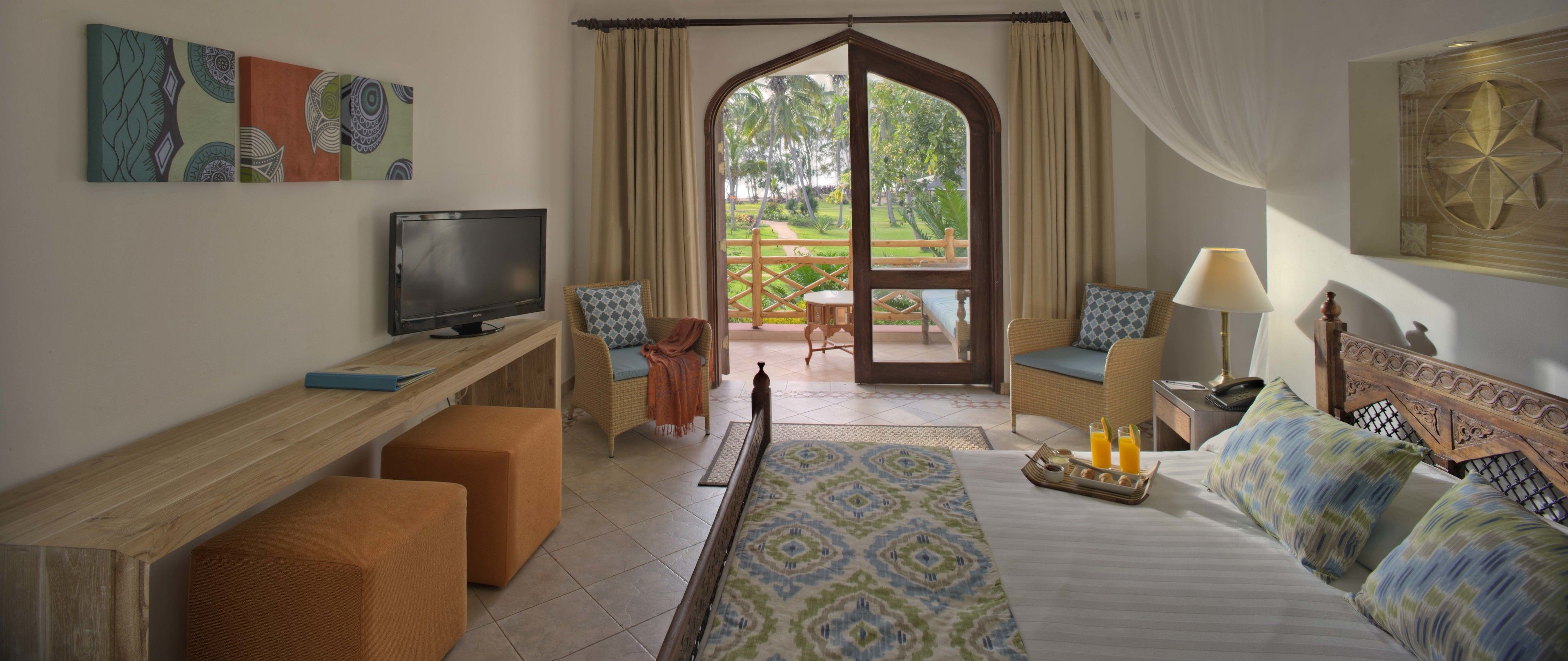 Habitación de hotel con una cama, un escritorio, televisión y vista a un jardín a través de un ventanal grande.