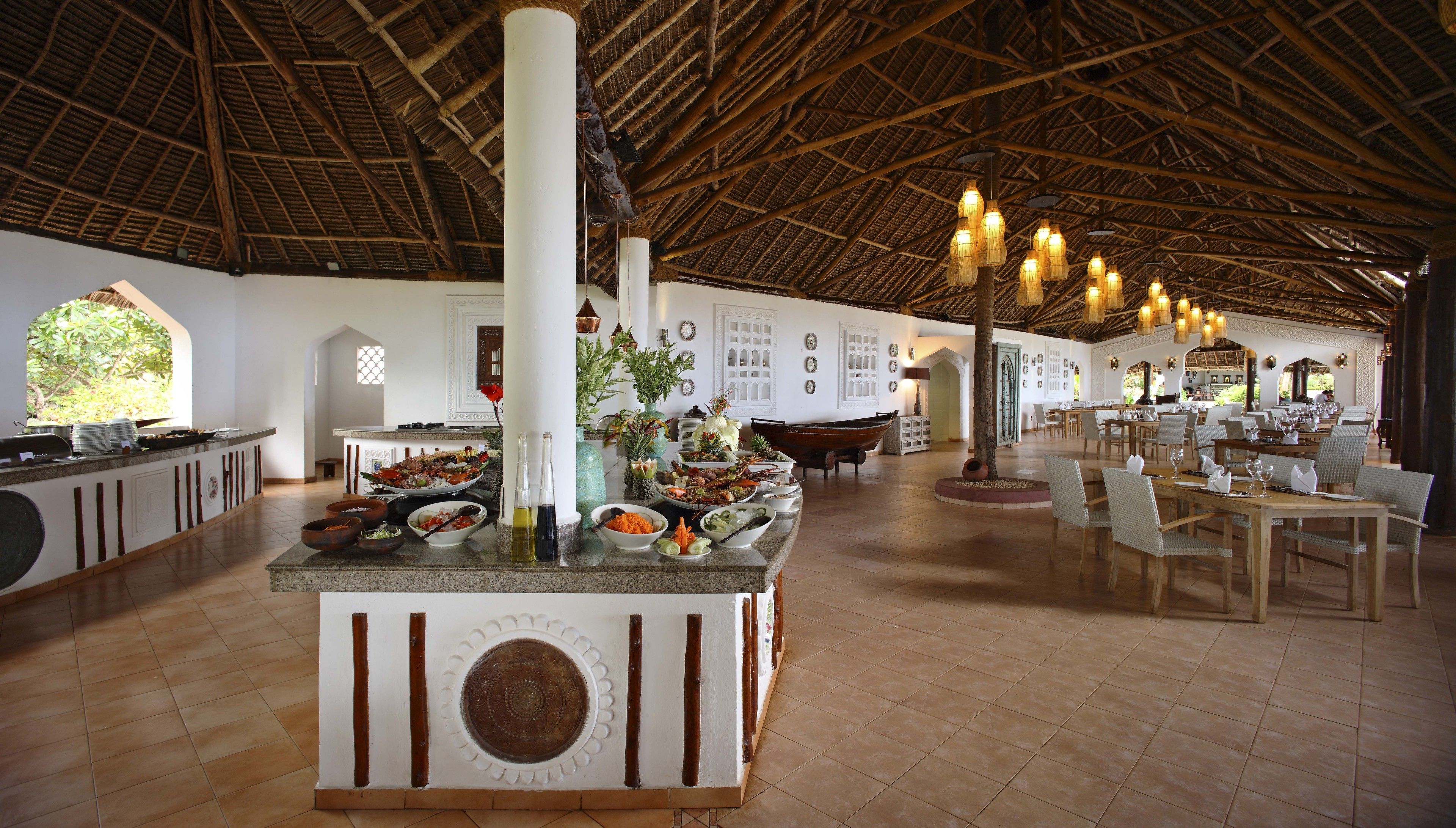 Een ruime binnenzone met een hoge kap en rieten dak, met een centraal buffet met diverse gerechten en een zithoek met tafels en stoelen.