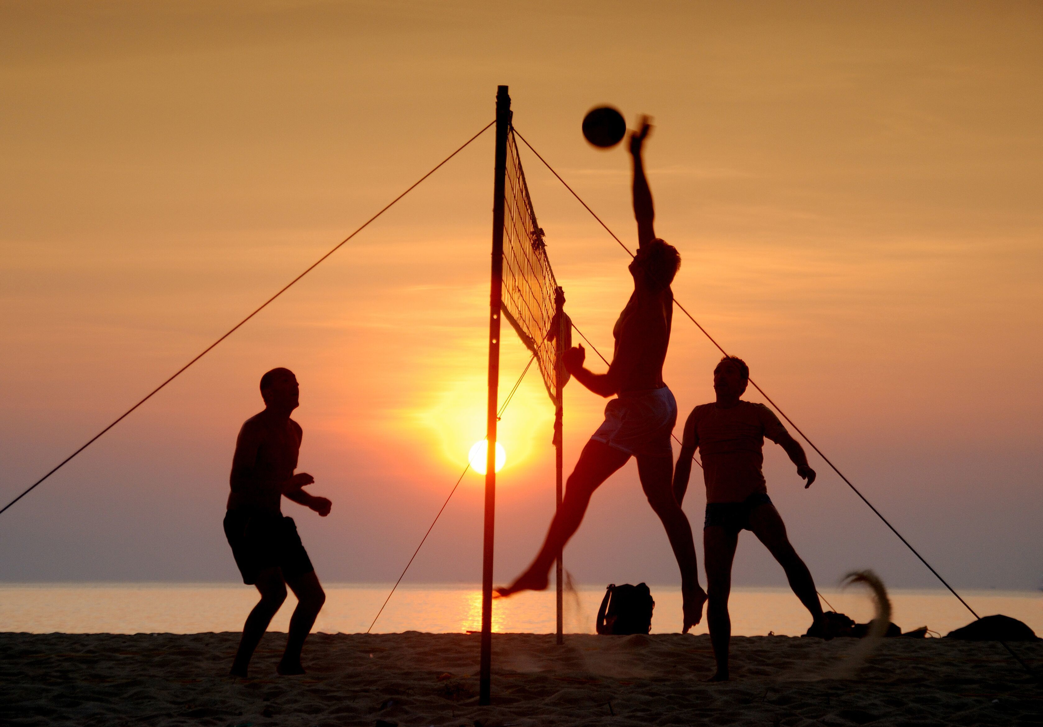 A beach volleyball game at sunset with a net and players silhouetted against the sky.