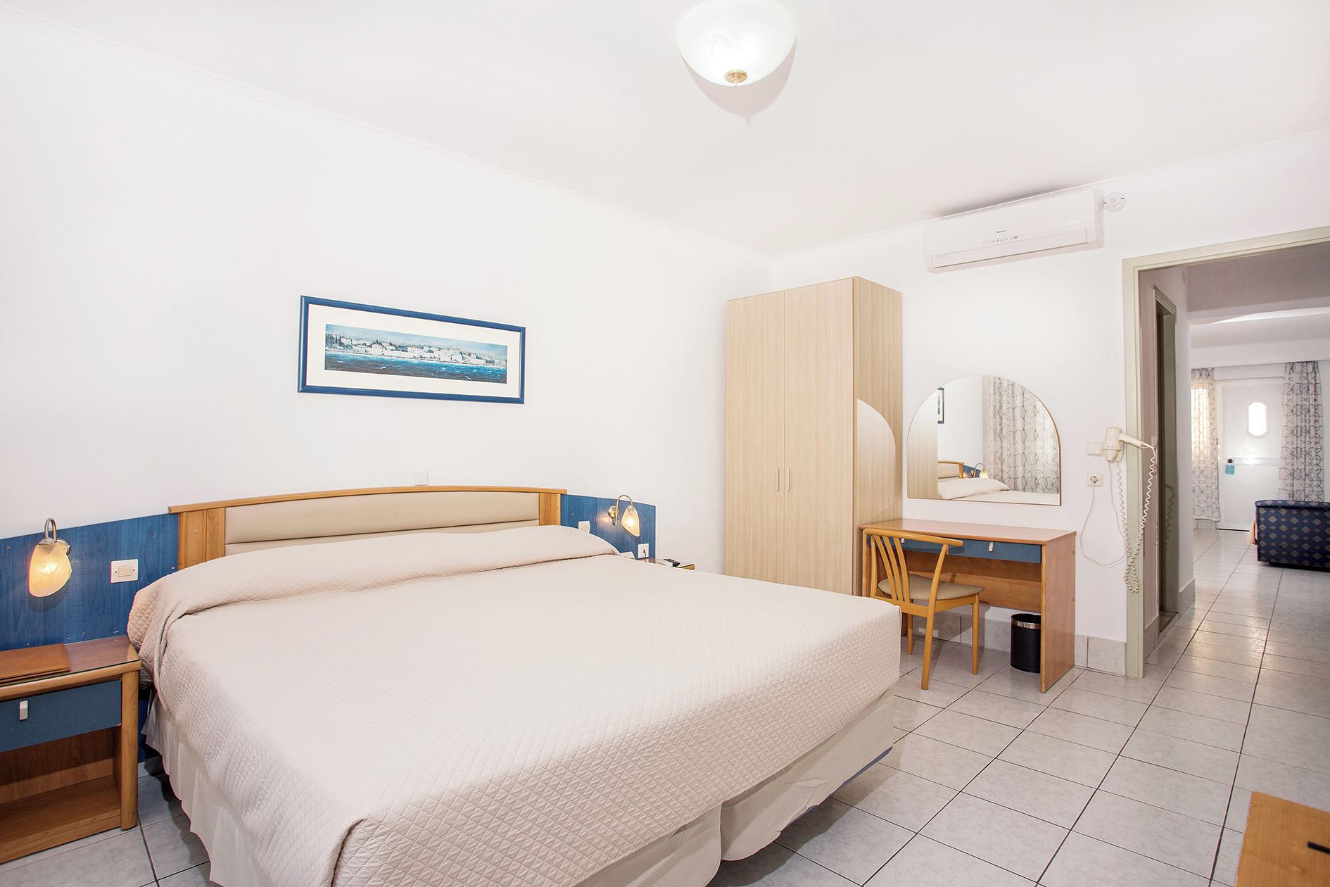 A hotel room with a bed, a desk, a wardrobe, a mirror, and a framed picture on the wall.