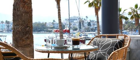 A wicker chair and table with a glass of water, a cup, and a glass of coffee on a sunny outdoor patio with palm trees and a view of the water and buildings in the background.