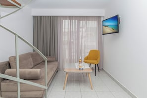 A modern living room with a sofa, a small table with a bottle, a chair, a TV mounted on the wall, and a staircase. - Kosta Palace (Kos)