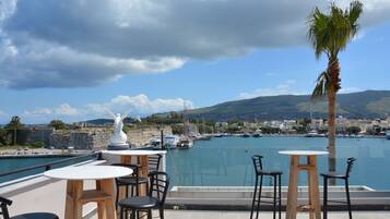 A terrace with a view of a marina, featuring a statue, wooden tables, and chairs.