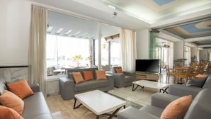 A modern living room with grey sofas, orange cushions, a flat-screen TV, and large windows offering a view of the outdoors.