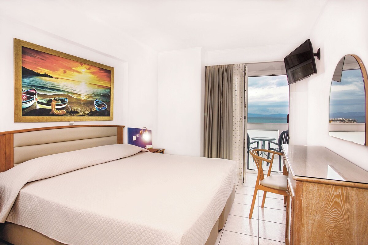 A hotel room with a bed, a desk, a chair, a TV mounted on the wall, and a painting of boats on the sea.