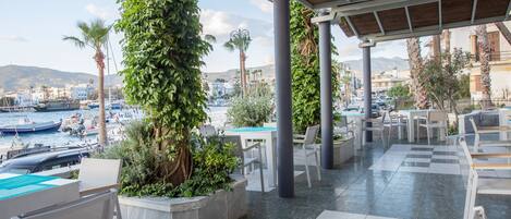 An outdoor seating area with tables and chairs, surrounded by greenery and overlooking a marina with boats.