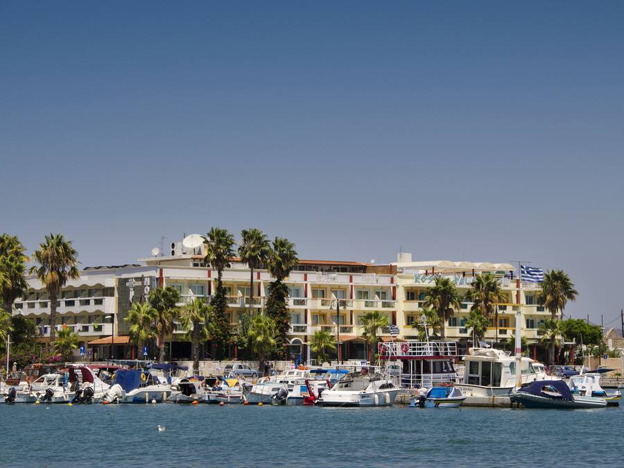A marina with multiple boats docked, a hotel with balconies, and palm trees in the background.