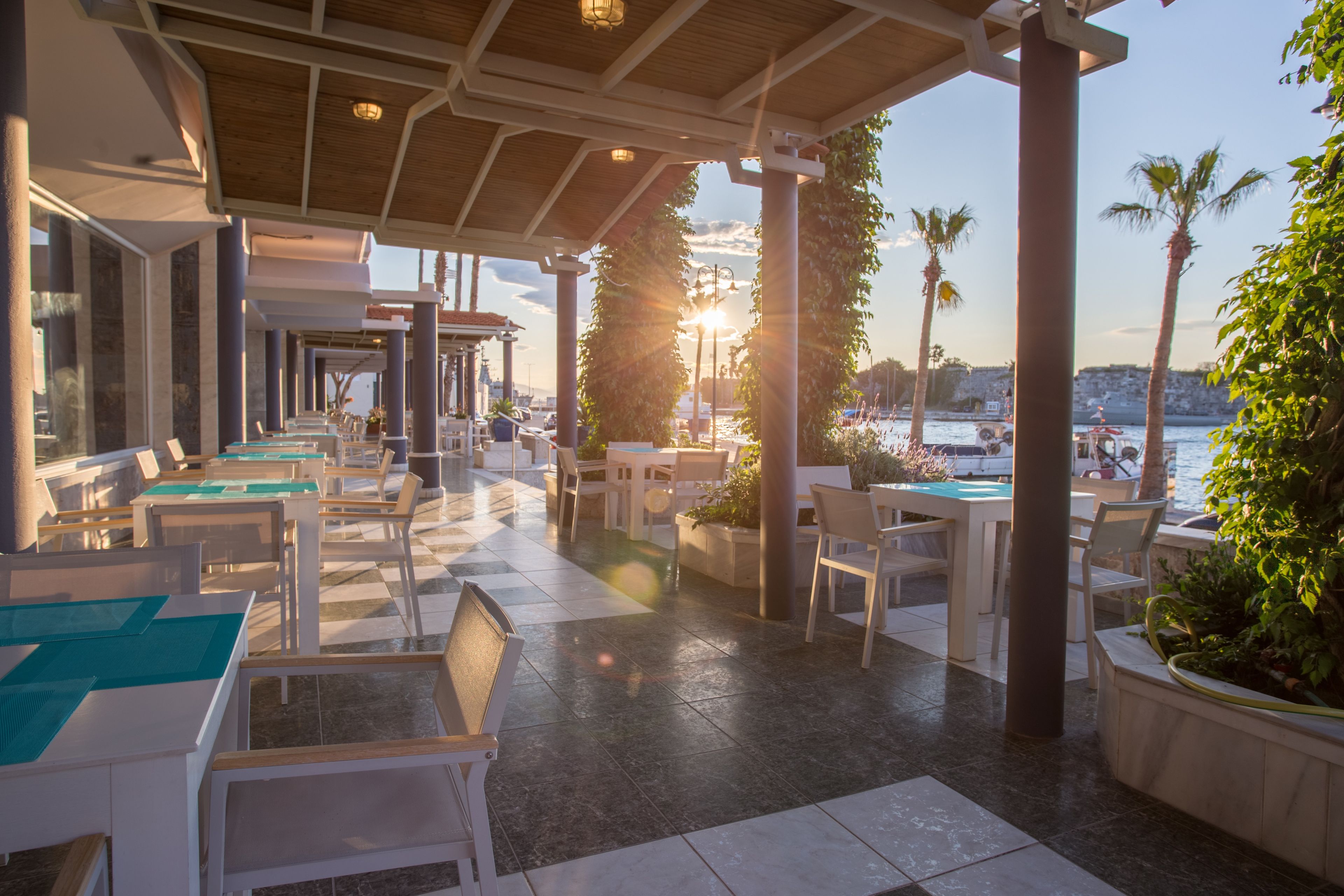 An outdoor seating area with tables and chairs, a view of the sea, and a sunset in the background.