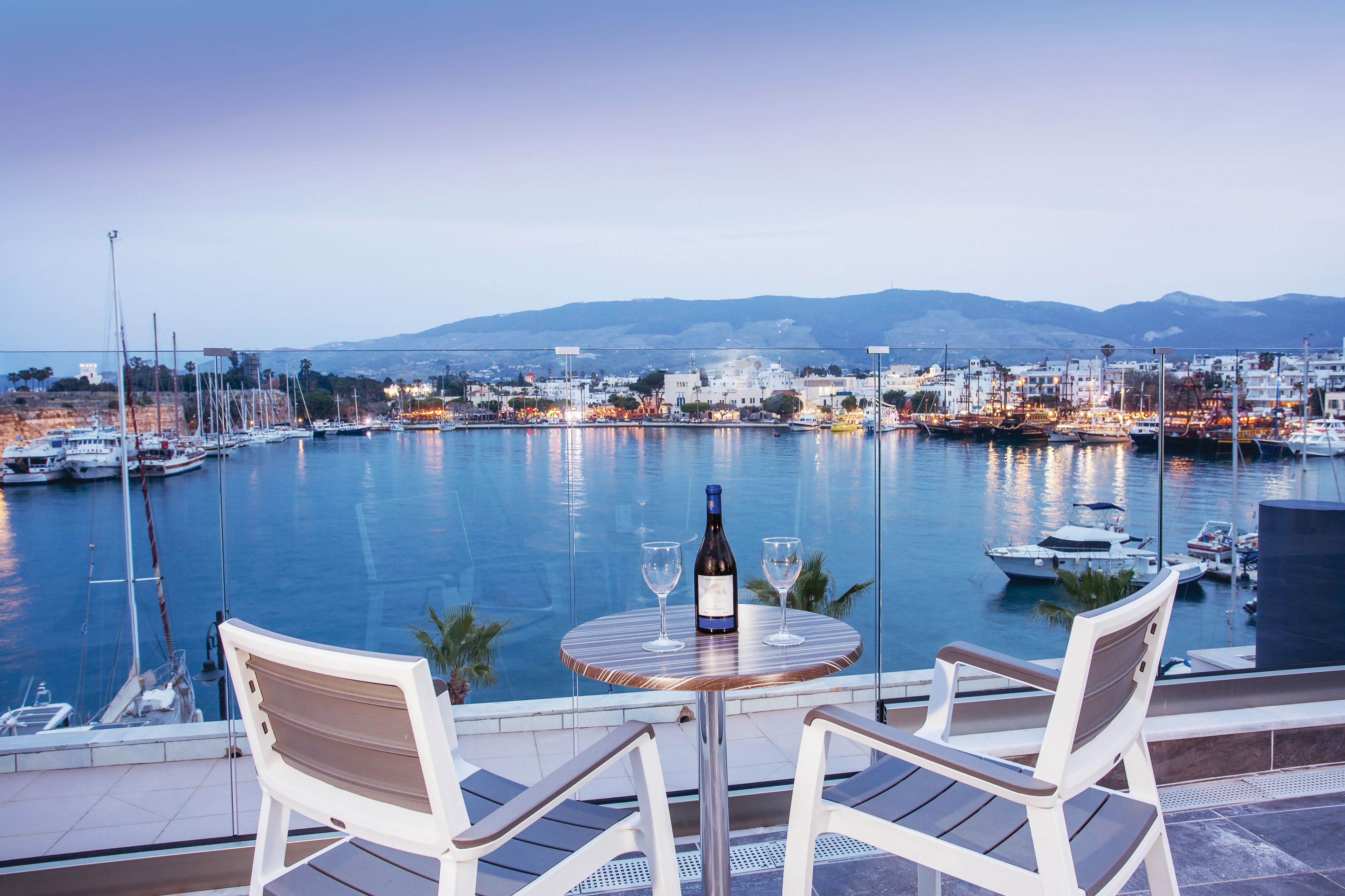 A table with two chairs and a bottle of wine, overlooking a marina with boats and a cityscape in the background.