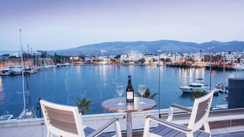A table with two chairs and a bottle of wine, overlooking a marina with boats and a cityscape in the background.