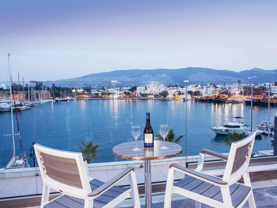 A table with two chairs and a bottle of wine, overlooking a marina with boats and a cityscape in the background.