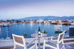 Una mesa con dos sillas y una botella de vino, con vista a un puerto deportivo con barcos y un paisaje urbano de fondo.