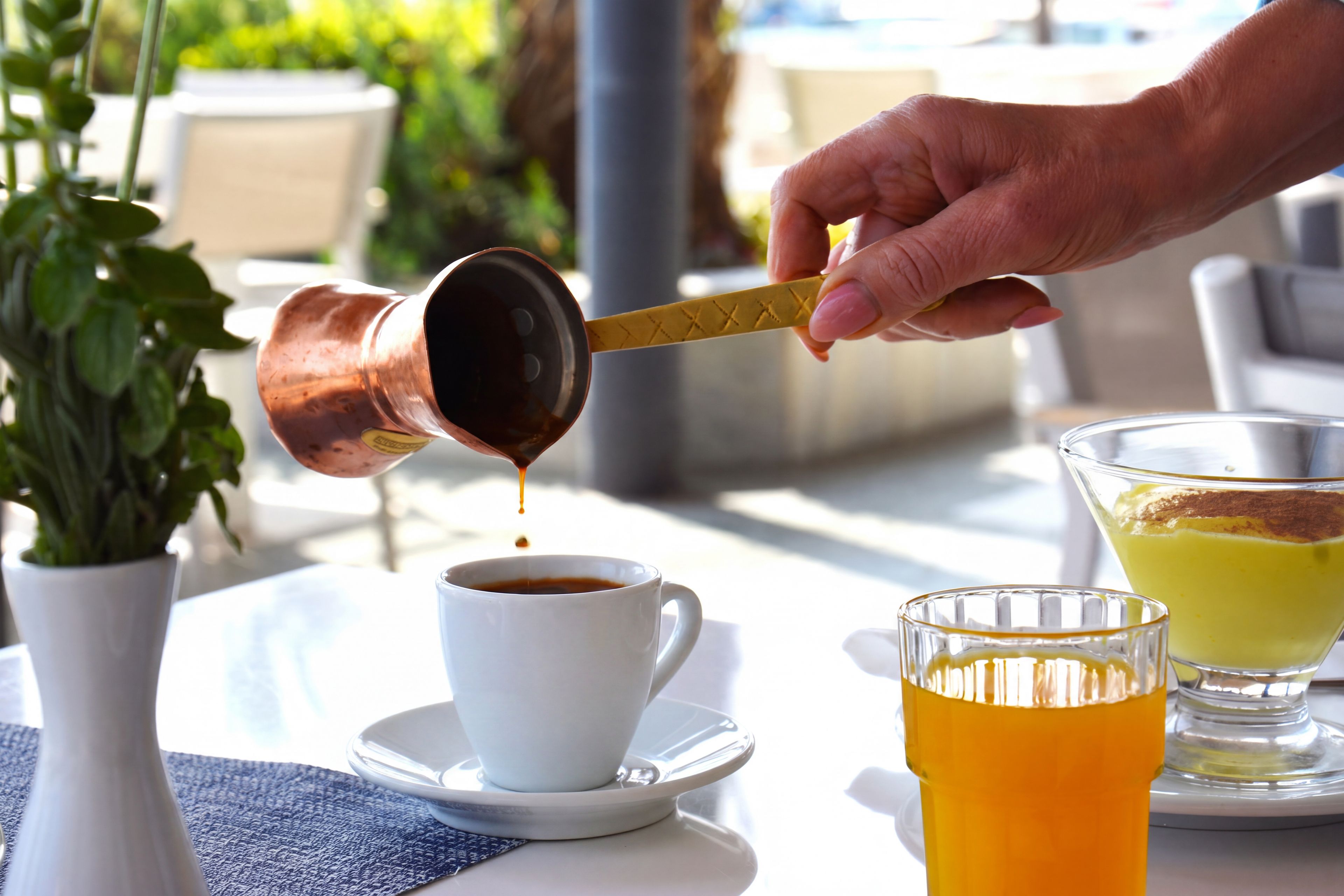 Eine Hand gießt eine dunkle Flüssigkeit in eine weiße Tasse, im Hintergrund steht ein Glas mit gelber Flüssigkeit und ein Blumentopf.