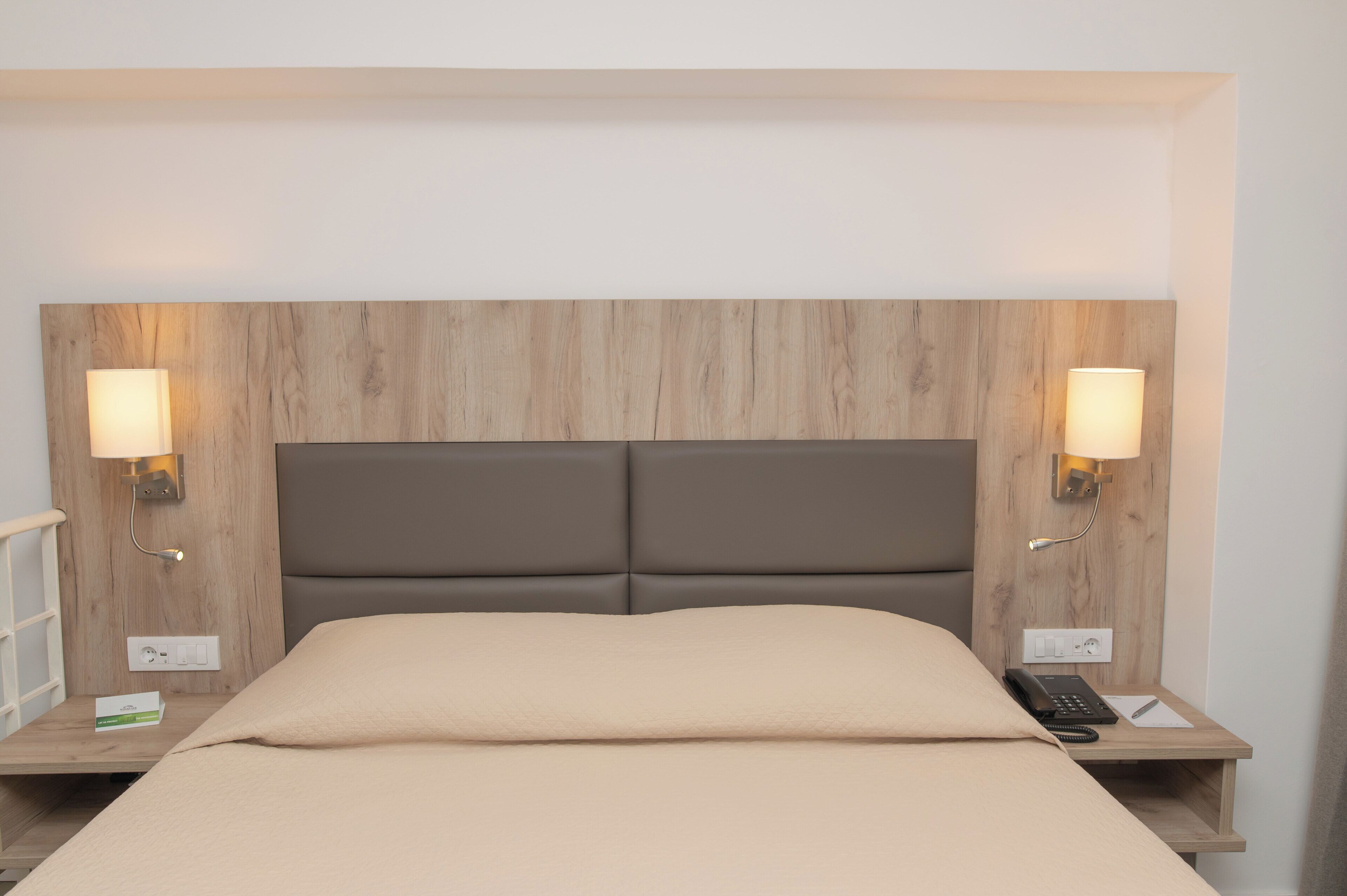 Habitación de hotel con cabecera de madera, dos lámparas de noche, cama con manta color beige, teléfono en la mesita de noche y una pequeña mesa con toma de corriente.
