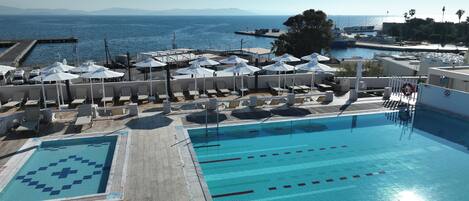 Una piscina con sillones de descanso y sombrillas, un muelle con barcos, y vistas al mar y a las montañas.