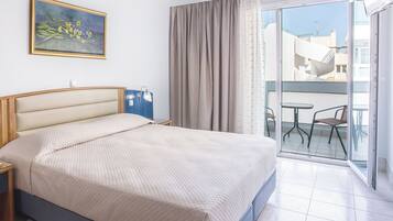 Ein Hotelzimmer mit Bett, Nachttisch, Lampe, einem Gemälde, einem Balkon mit Sitzgelegenheiten und Blick auf Gebäude.