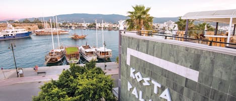 Un puerto deportivo con barcos amarrados, un edificio con el rótulo "KOSTA PALACE" y vistas al agua y colinas distantes.