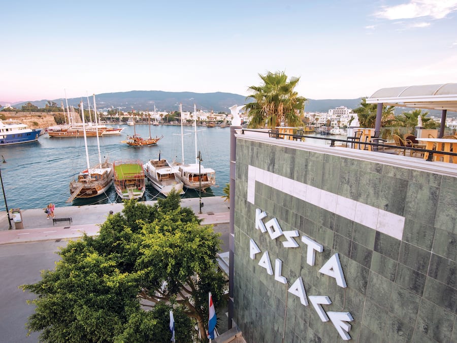 Un puerto deportivo con barcos amarrados, un edificio con el rótulo "KOSTA PALACE" y vistas al agua y colinas distantes.