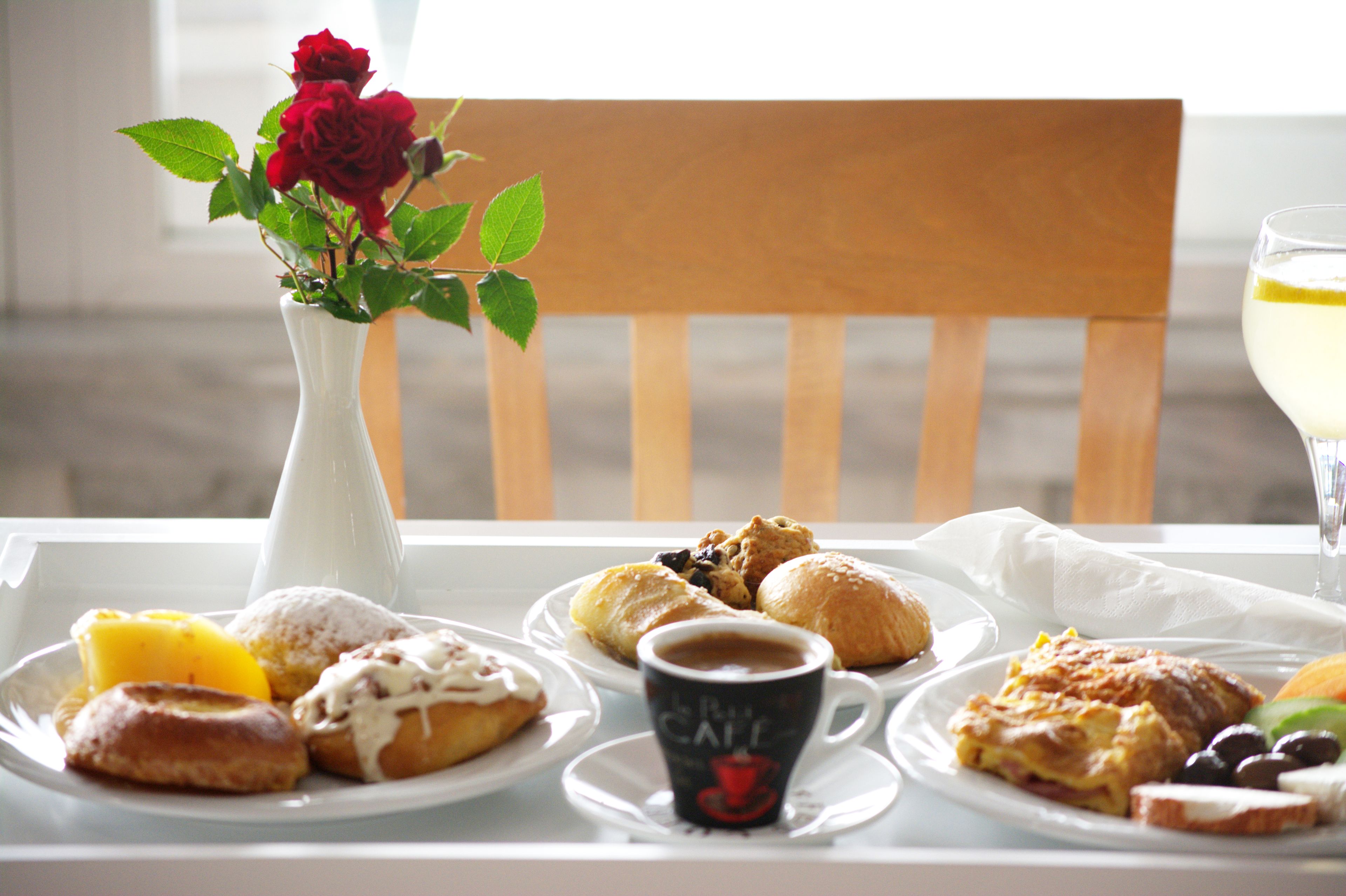 A table with a variety of pastries, a cup of coffee, and a glass of white beverage, with a vase of flowers and a wooden chair in the background.
