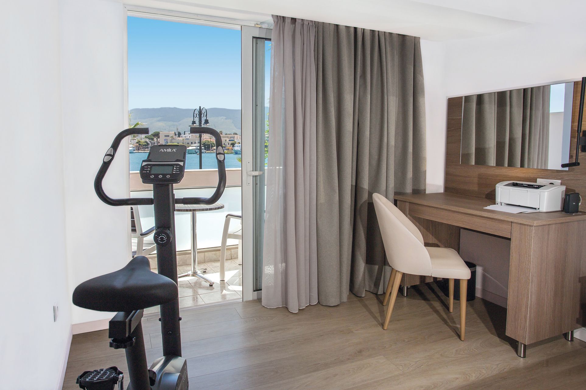 Quarto de hotel com escrivaninha, cadeira, bicicleta ergométrica e vista para o mar.