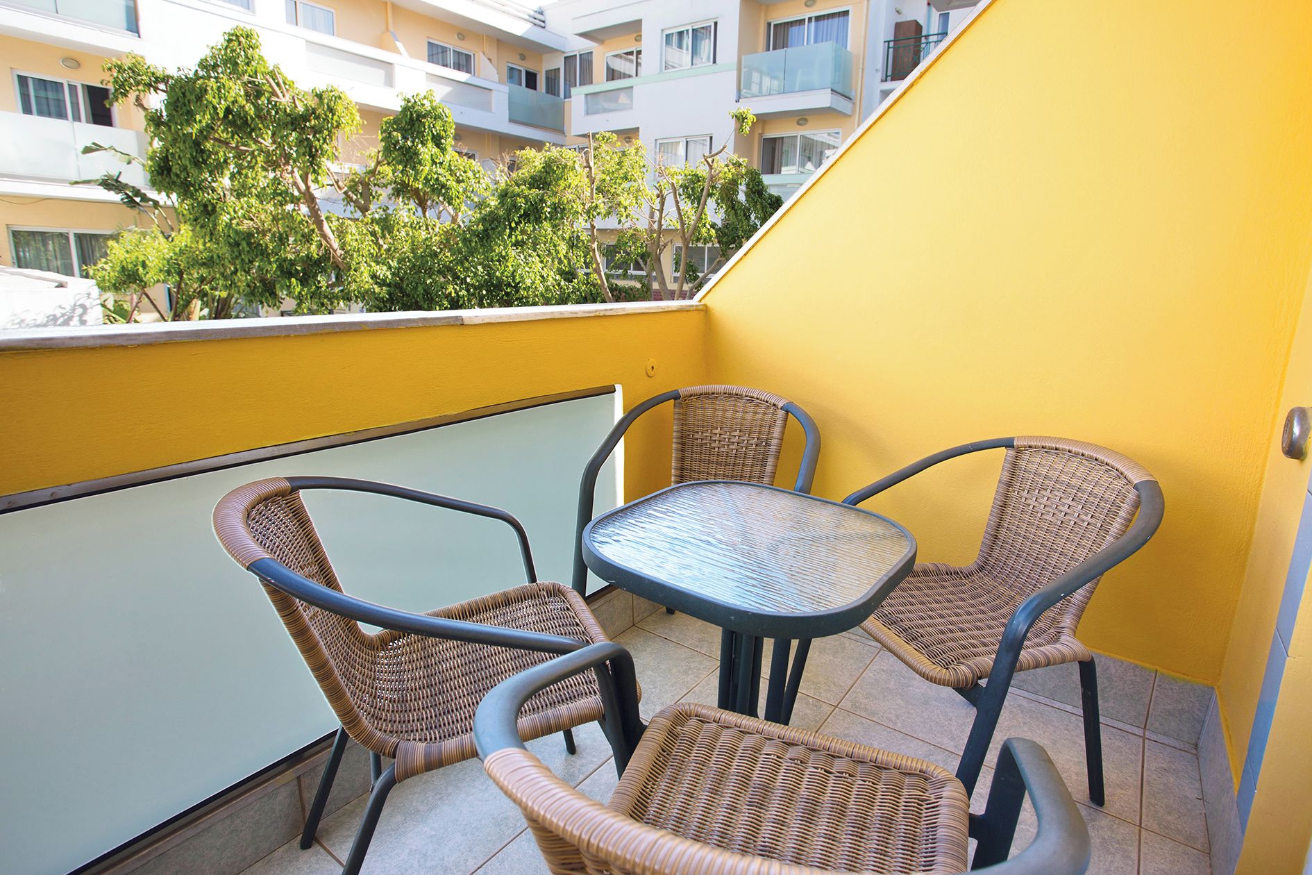 A balcony with wicker chairs and a table, overlooking a yellow building and trees.